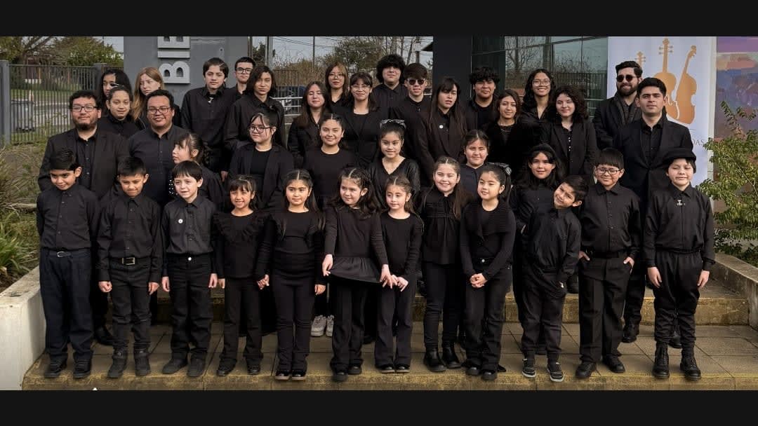 Los integrantes de la Orquesta de Cuerdas Infantil Juvenil de Frutillar, niños y jóvenes vestidos de negro, posan frente a la Biblioteca Municipal