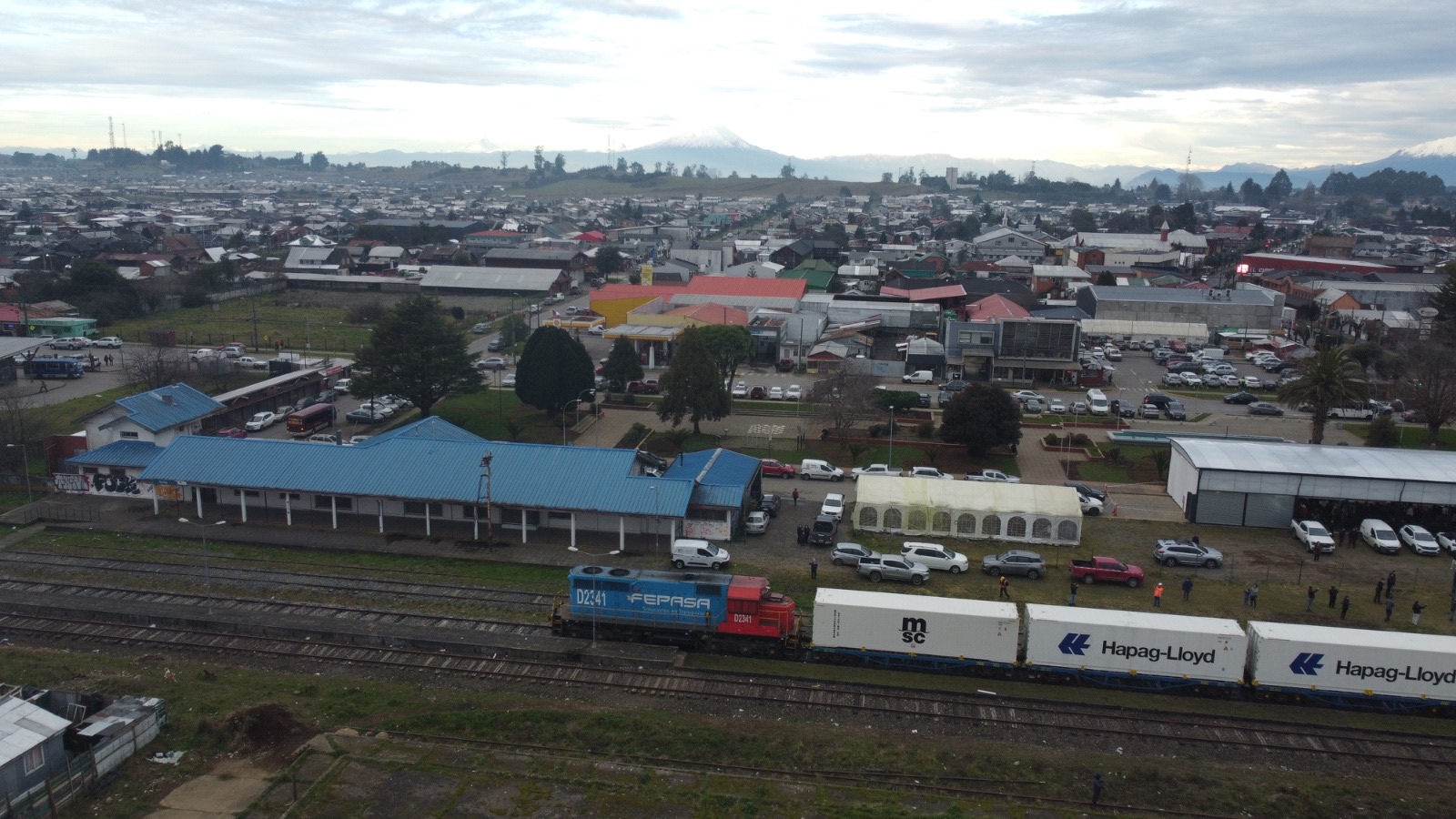 Vista aérea del corredor ferroviario de Frutillar con un tren de contenedores junto a la antigua estación, el pueblo y el volcán Osorno al fondo