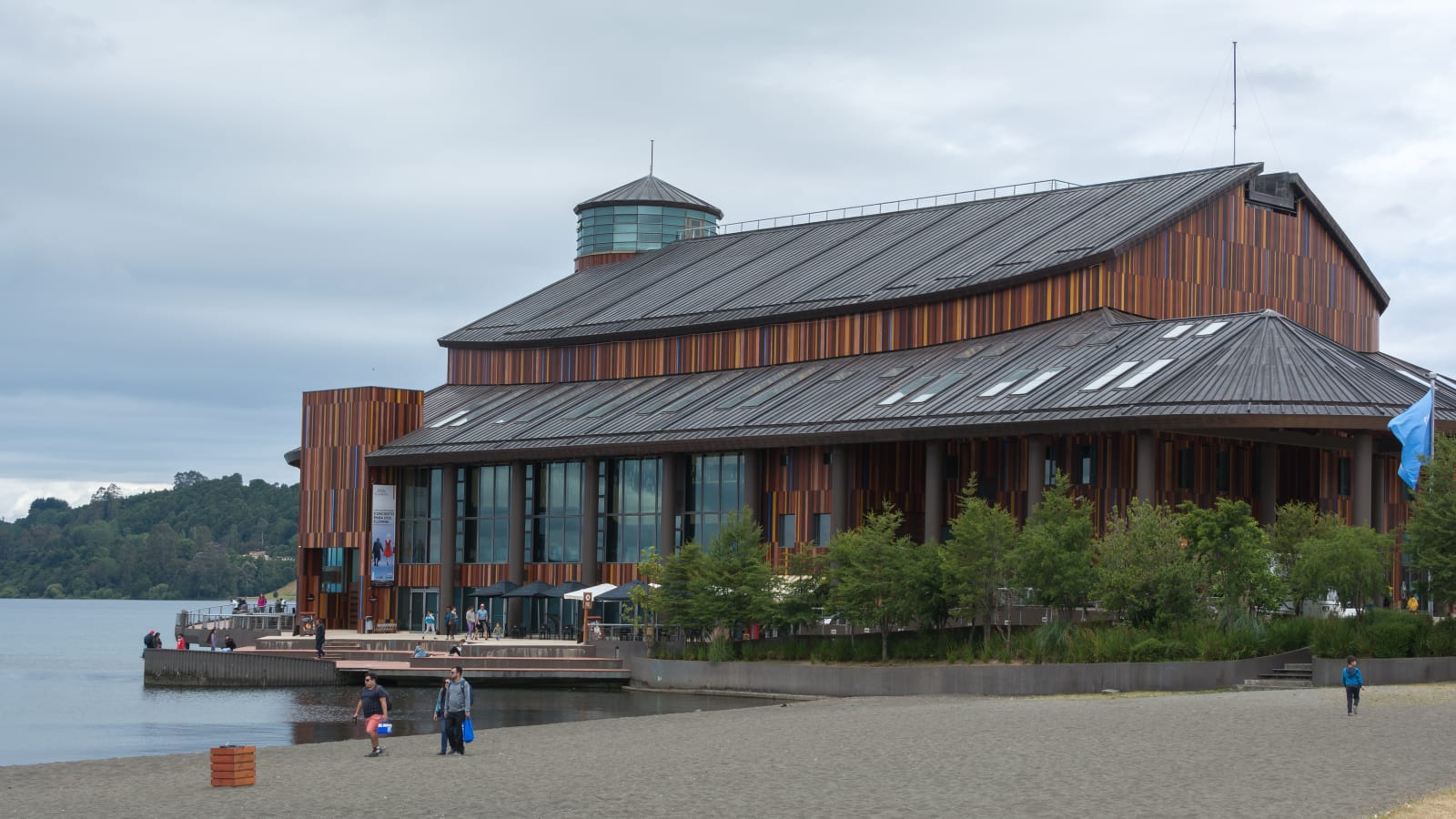 Fachada de madera del Teatro del Lago de Frutillar vista desde la playa, con personas caminando frente al edificio