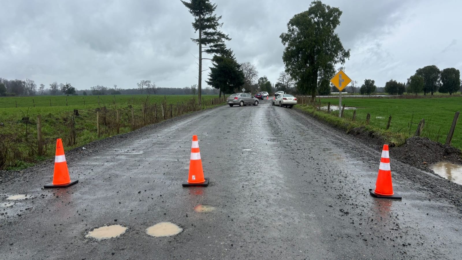 Conos naranjos cortando un camino rural mojado entre praderas, con vehículos detenidos al fondo, entre Casma y Copiwel en Frutillar