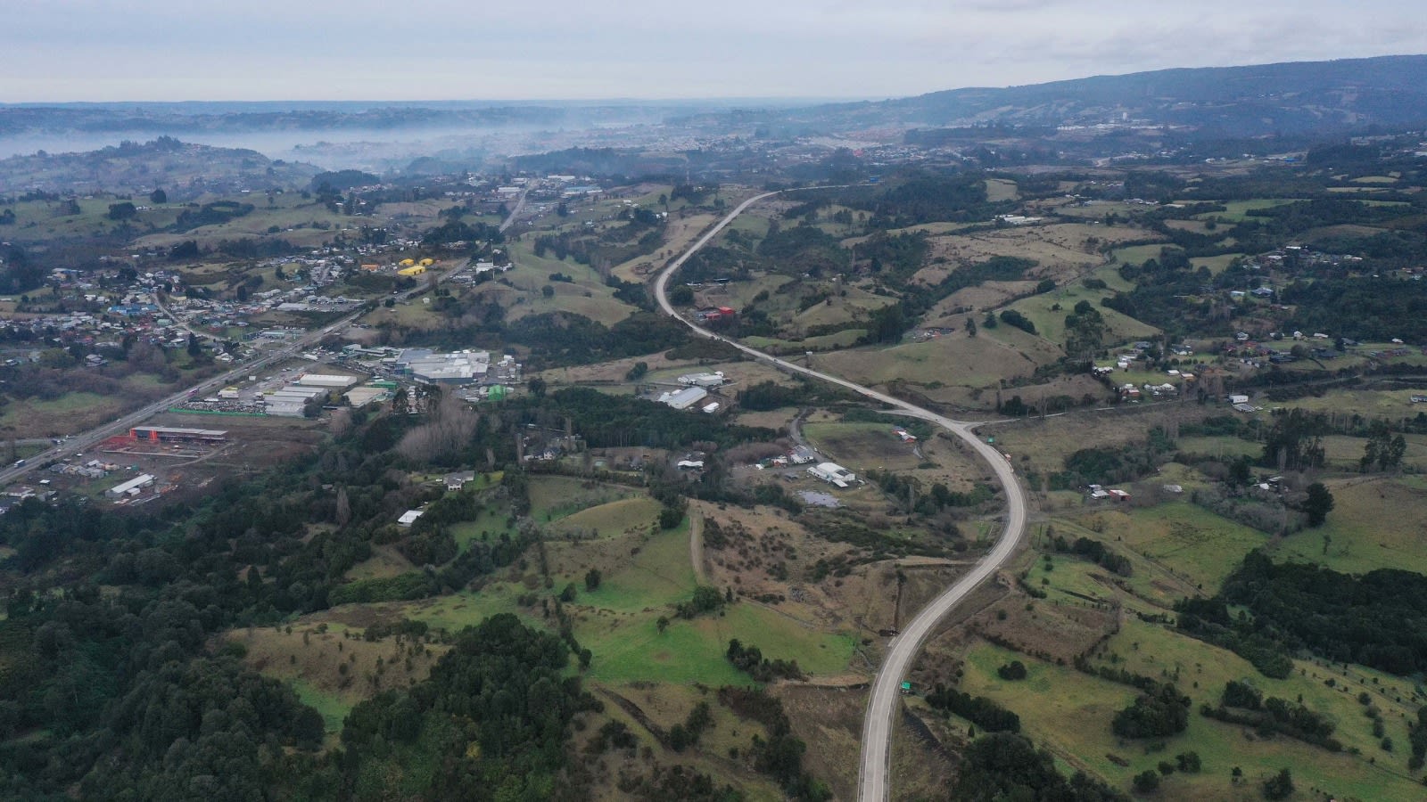Vista aérea del trazado del bypass entre Castro y Chonchi cruzando campos y bosques de Chiloé, con la ciudad al fondo