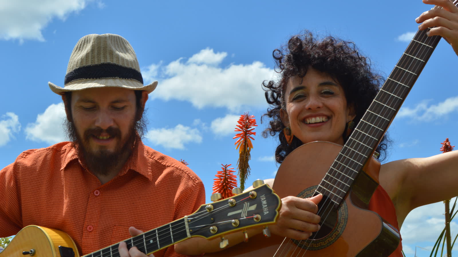 Dos músicos sonríen mientras tocan guitarras al aire libre, bajo un cielo azul