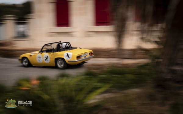 Straw bales lining the circuit as a classic car passes the Mdina city gate