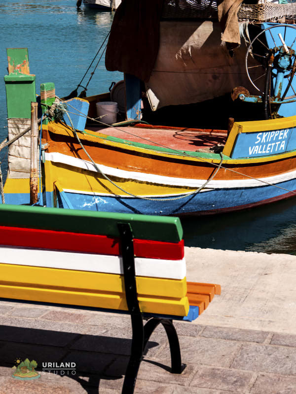 A bench painted in luzzu colours with a fishing boat in the background