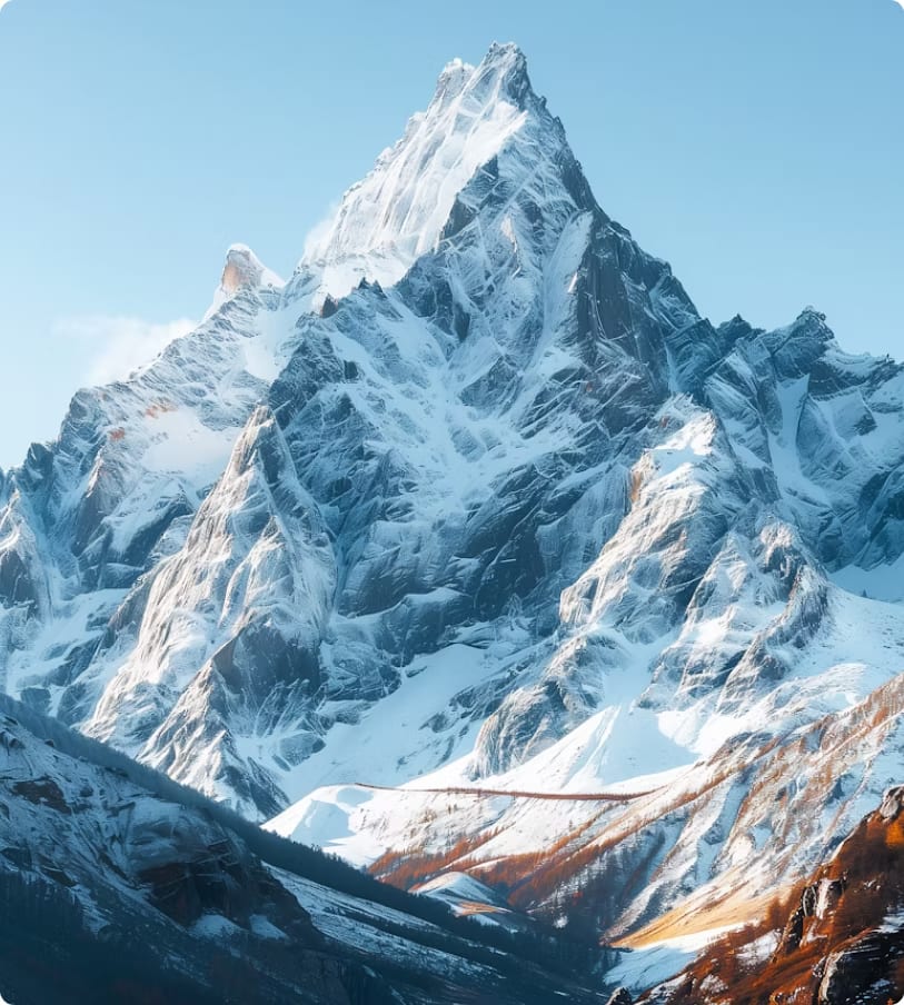 Snow-covered towering mountain peak under clear blue sky with some autumn trees at the base.