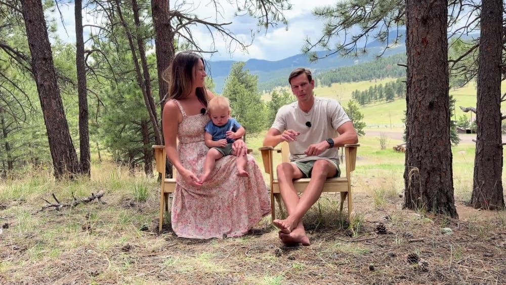 Ashley and Brian seated side by side in a mountain meadow, the baby on her lap