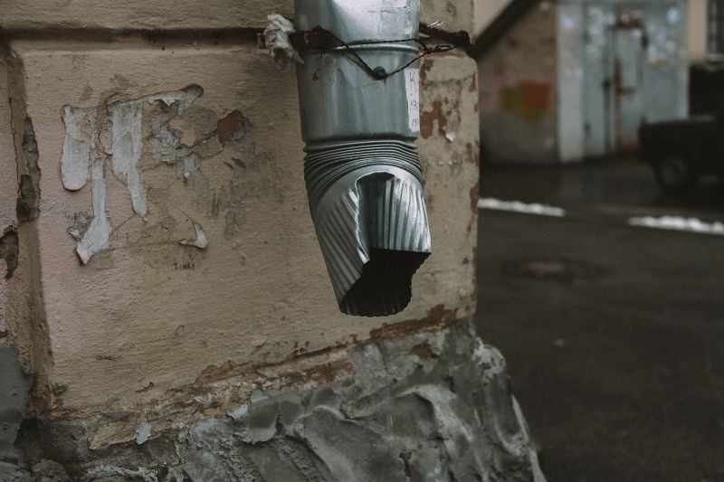 Close-up of a gray downspout attached to a weathered wall above a stone base.