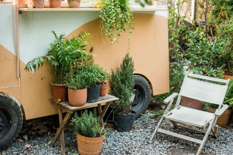 Potted plants on a table beside a beige trailer, with a white folding chair on gravel.
