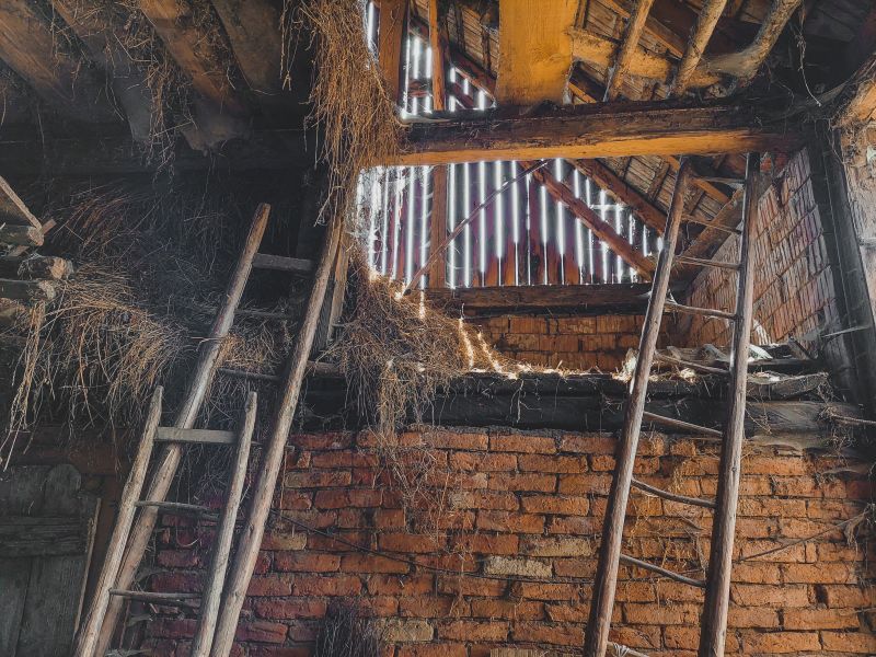 Inside a damaged brick attic with a ladder, exposed beams, straw insulation, and a slatted window opening