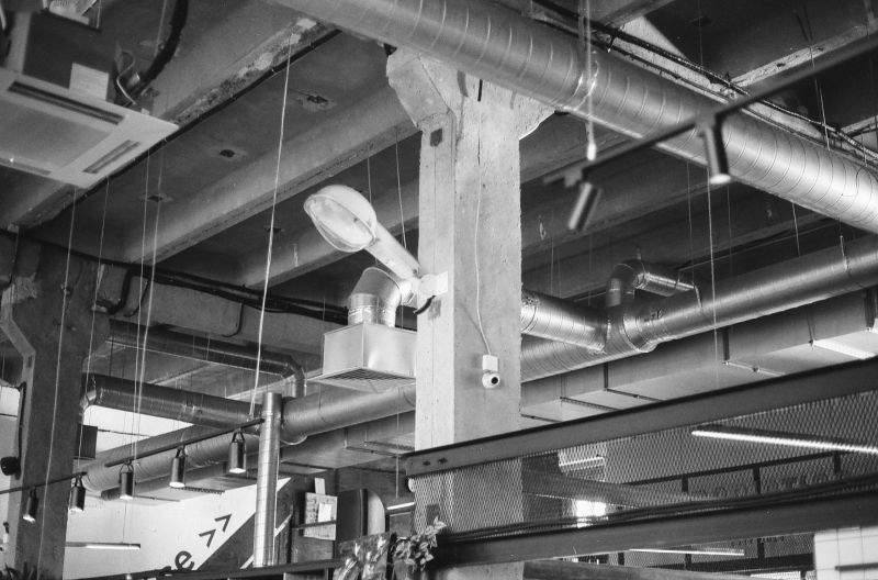 Black-and-white industrial ceiling with exposed ducts, pipes, beams, and hanging lights in a warehouse interior