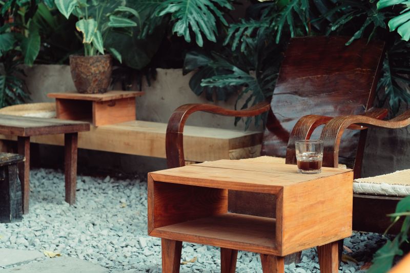 Outdoor wooden chairs and tables on gravel, with lush green plants in the background.