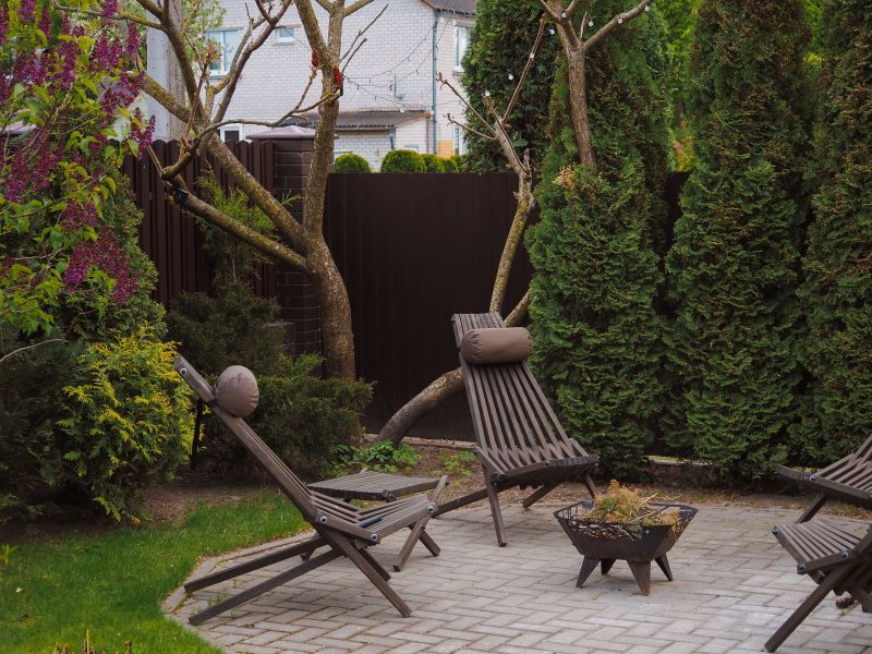 Garden patio with two wooden lounge chairs, a fire pit, and tall hedges around a stone patio