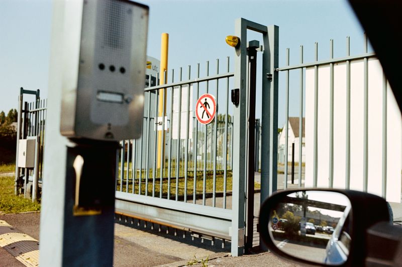 Metal gate and fence beside a road, viewed from a car with side mirror visible.