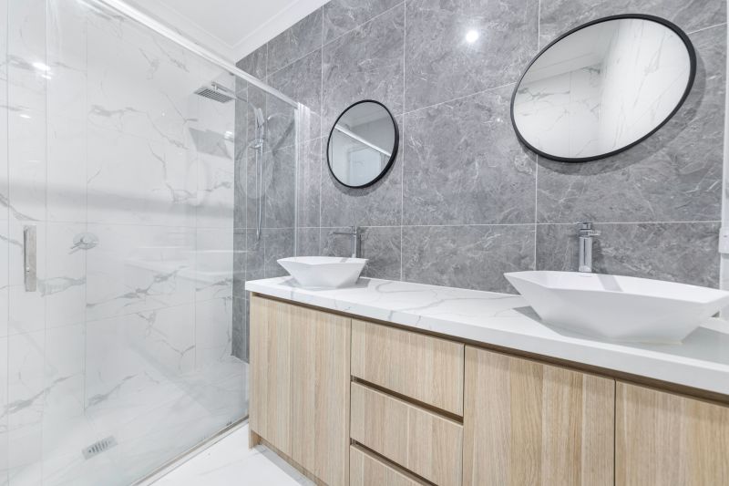 Modern bathroom with double sinks, gray tile walls, and a large oval mirror