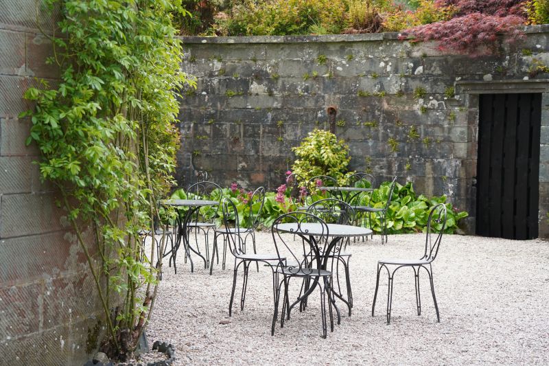Outdoor patio with black metal tables and chairs on white gravel beside a stone wall and greenery