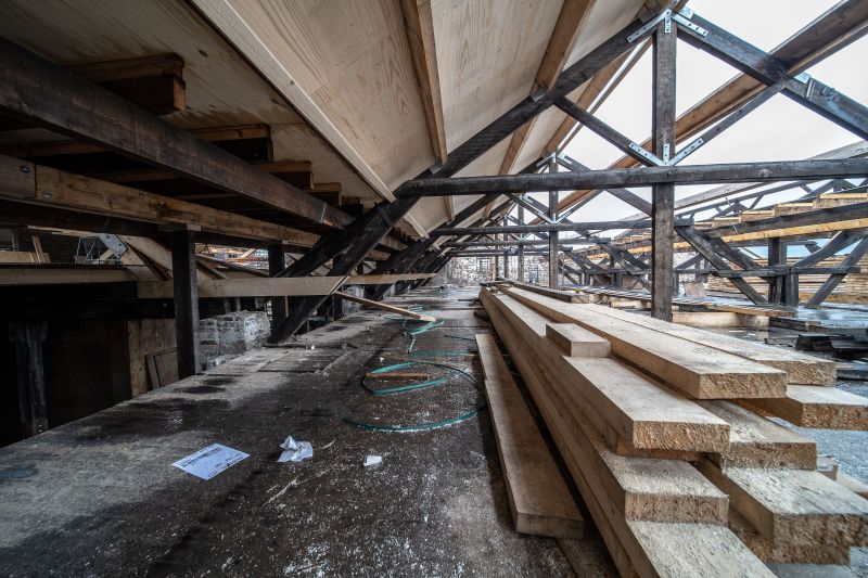 Abandoned industrial warehouse with wooden beams, broken roof trusses, and stacked lumber on a concrete floor