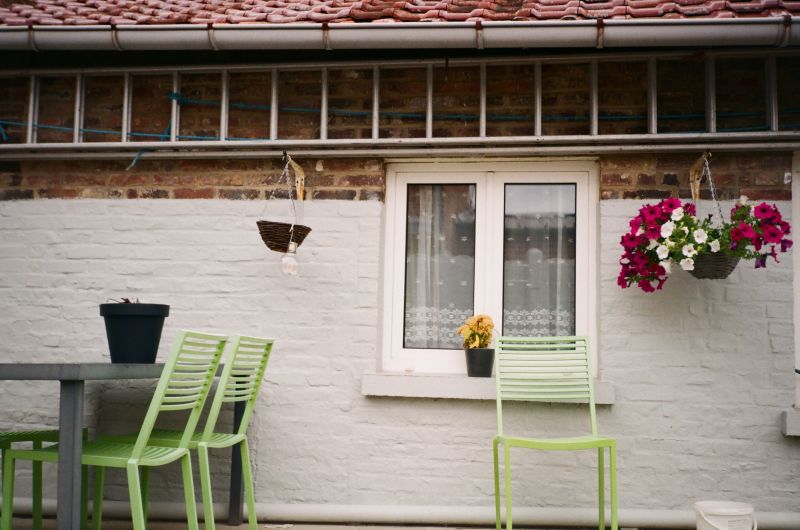 White patio with lime-green chairs, table, window, and hanging pink flowers on a brick wall.