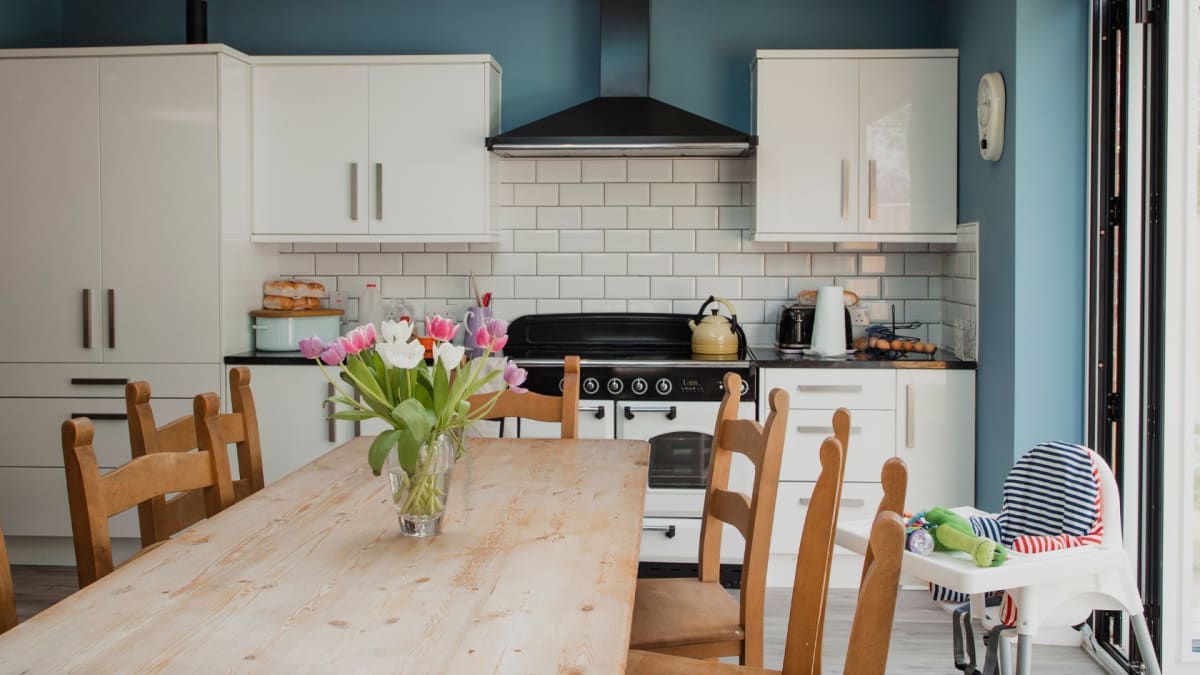 Kitchen with white cabinets, wooden table and chairs, blue wall, flowers.