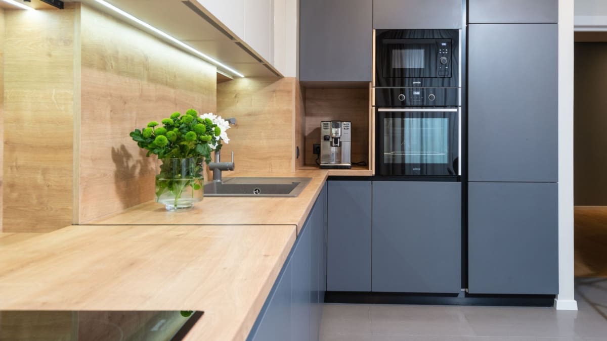 Modern kitchen with gray cabinets, light wood countertops, built-in oven and microwave, and a coffee machine.