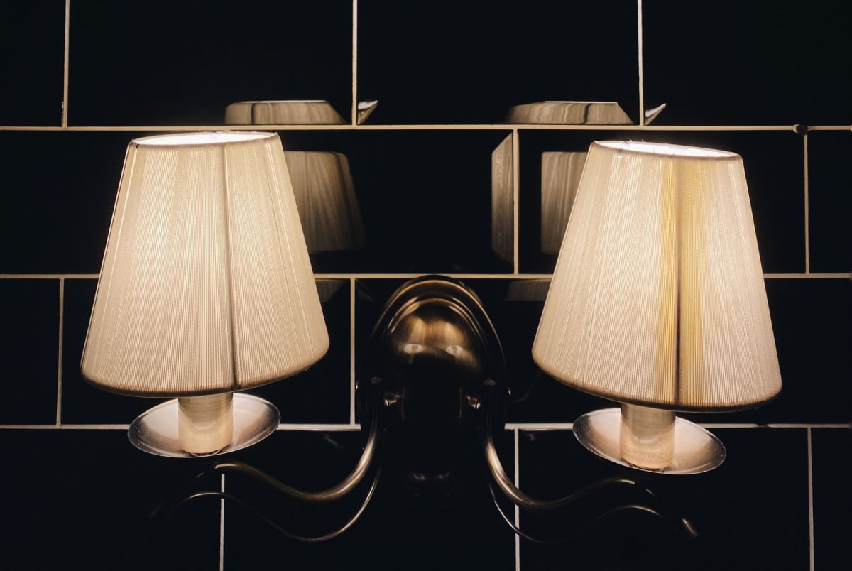 Pair of cream pleated wall sconces on a dark tiled wall, with a central brass fixture between them.