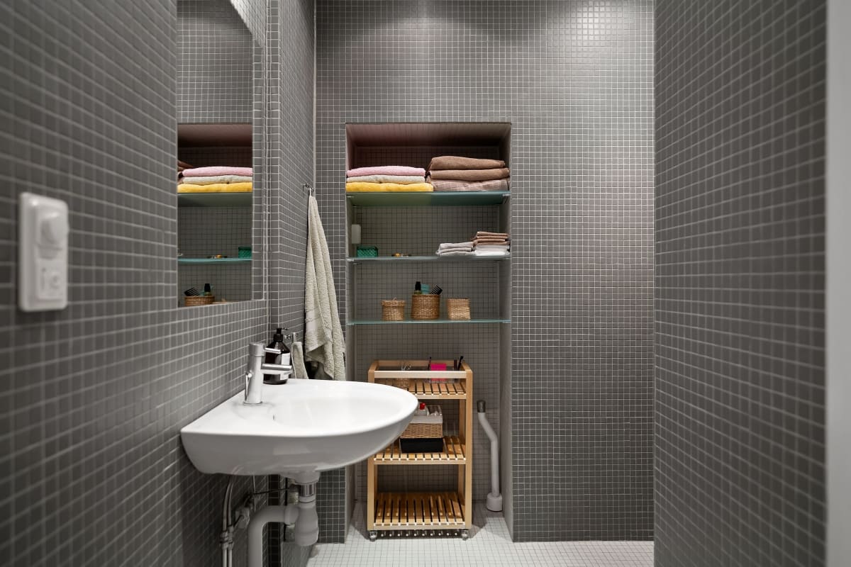 Small tiled bathroom with a white sink, open storage shelves, folded towels, and a wicker hamper.