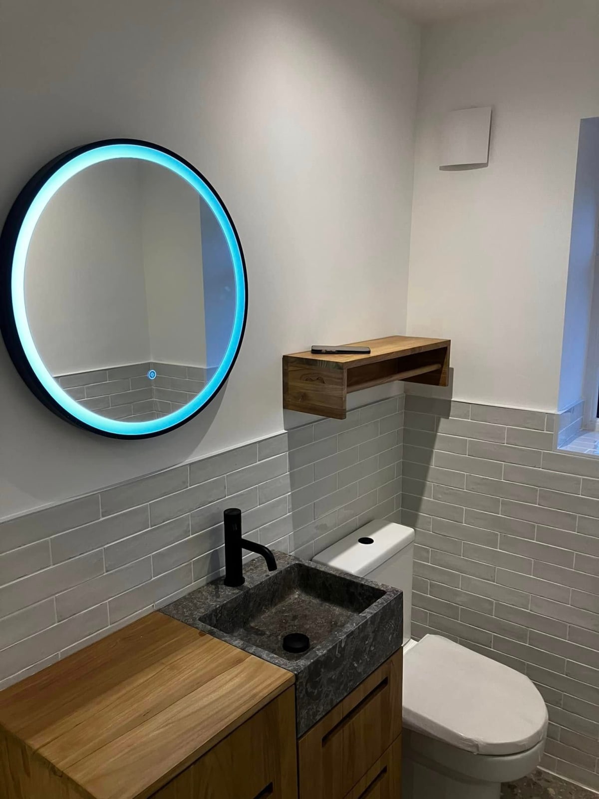Modern bathroom: round mirror with blue light, wooden vanity, black faucet, and toilet.