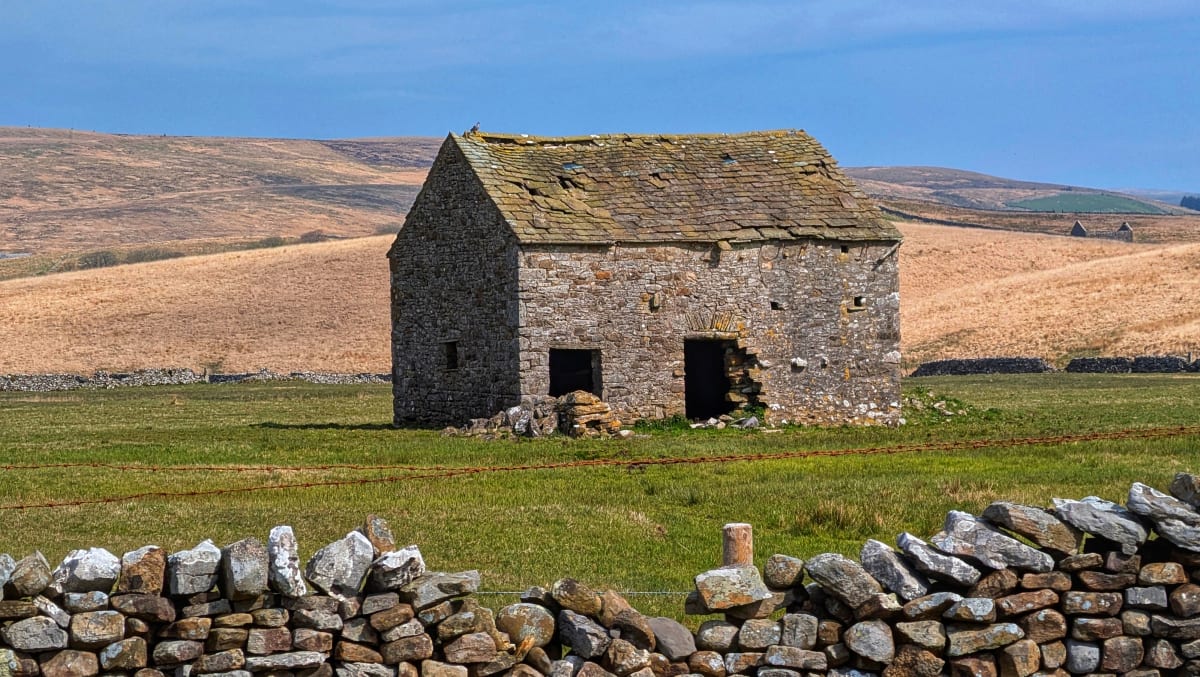 Small stone cottage in a grassy field, with dry hills in the background and a stone wall in front.