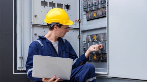 Technician in yellow hard hat inspecting an electrical control panel, holding a laptop in an industrial setting