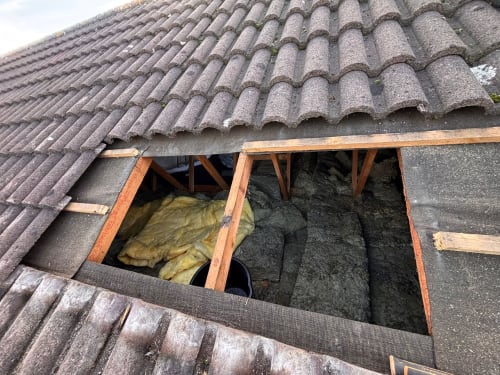 A section of roof tiles removed to reveal wooden rafters and yellow fiberglass insulation inside an attic.