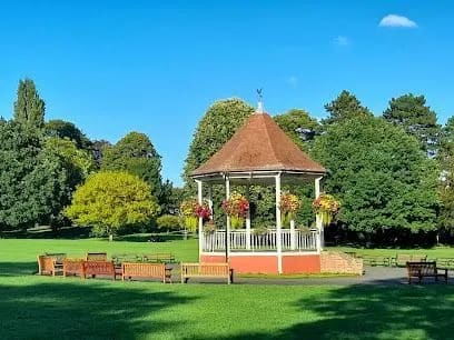 Bandstand in a park on a sunny day with green grass, trees, and hanging flower baskets.