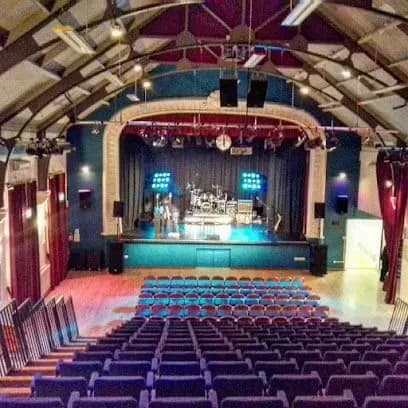 Auditorium interior with stage, rows of blue seats, stage with band setup, and red curtains.