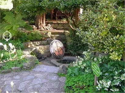 Stone garden path leading to a well with an ornamental object hanging above, surrounded by plants and trees.