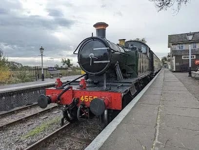 Steam train on a platform, numbered 4555. Black, green and red colors. Cloudy day, station setting.