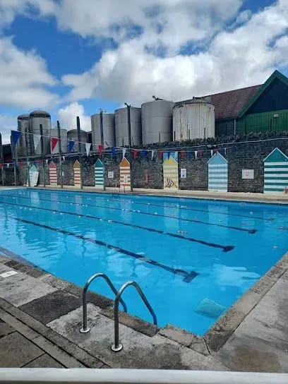 Outdoor swimming pool with blue water and lane markers, surrounded by colorful changing sheds and tanks under a cloudy sky.