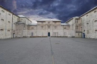 Exterior view of a large, stone prison building with an empty courtyard. Dark, stormy sky overhead.