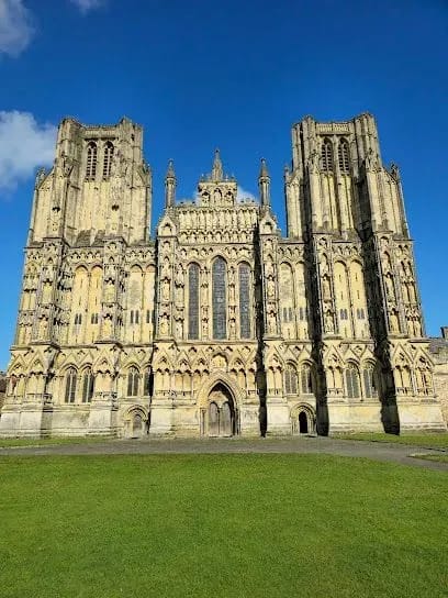 Wells Cathedral facade with two towers on a green lawn under a clear blue sky.