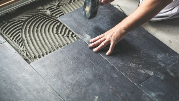 Person laying dark gray floor tiles with a hammer and mortar in a construction setting.