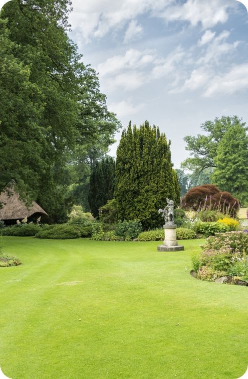 Lush green lawn with a statue, tall green trees, and a thatched roof building under a cloudy sky.