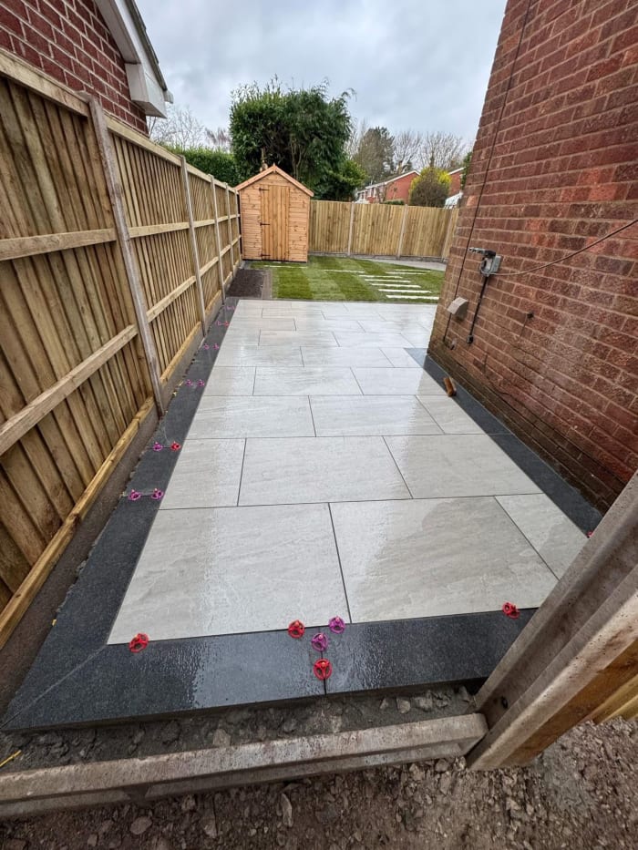 Newly paved patio with black border, next to wooden fence and brick wall. Small shed in the background.