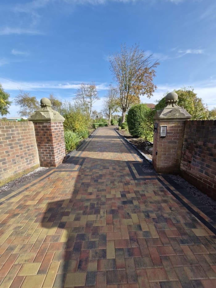 Brick pathway lined with pillars under a blue sky, leading towards trees and greenery.