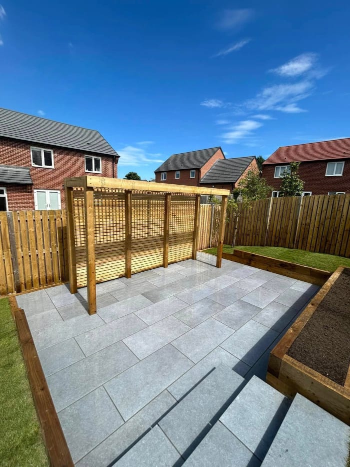 Wooden pergola on a gray paved patio in a backyard with a fence and houses under a blue sky.