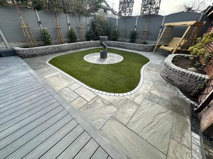 A modern garden with stone paving, a small lawn, and a sculpture, surrounded by a low wall and wooden fence.