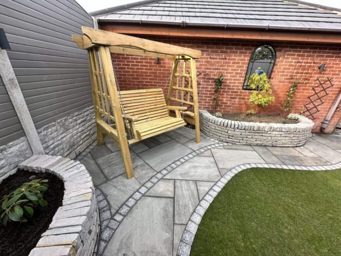 Wooden swing seat in a paved garden, near a brick building and a curved stone bed.