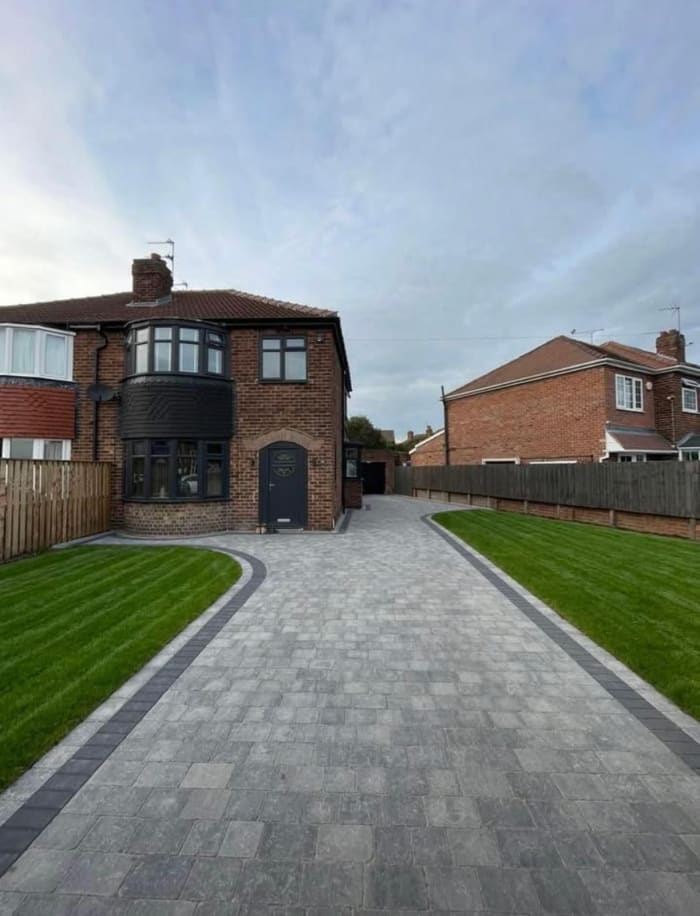 Brick house with grey driveway, green lawn, dark door and windows. Cloudy sky.