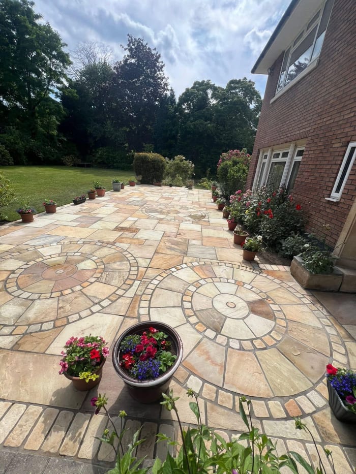 Stone patio with mosaic pattern, bordered by potted flowers, next to a brick building.