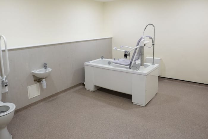 Accessible bathroom with bathtub-style chair lift, wall-mounted sink, and toilet; beige and white colors.