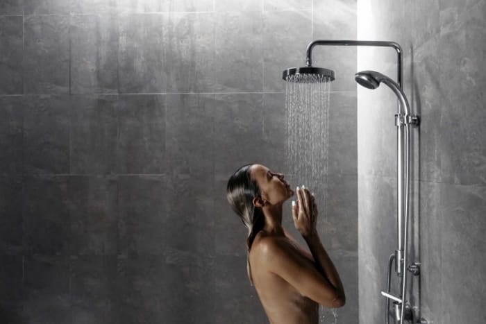 Woman in shower, head tilted back, arms raised to face, water streaming down. Gray tiled walls.