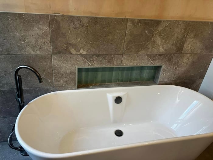 White bathtub with black faucet, in front of gray tile wall with inset shelf.