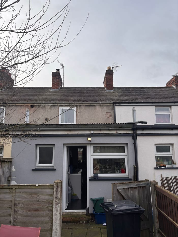 A grey, single-story rear house extension with a doorway and window, attached to a traditional two-story brick row house.