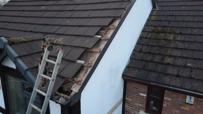 A ladder leans against a residential roof with missing tiles and exposed debris along the edge of a white wall.